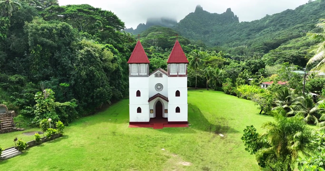 La Chiesa di Elisa si erge con due campanili rossi gemelli su un prato curato a Tautira, Tahiti, Polinesia Francese; montagne nebbiose della giungla e palme circondano il terreno della cappella bianca. Vista aerea con drone.