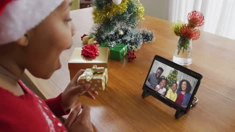 Mujer-Afroamericana-Con-Gorro-De-Papá-Noel-Usando-Tableta-Para-Videollamada-Navideña,-Con-Familia-En-Pantalla