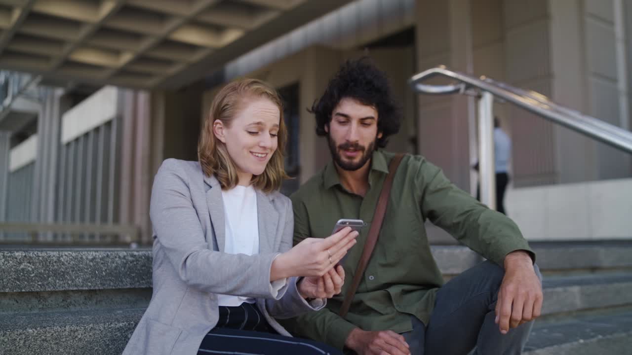 una atractiva mujer bien vestida en ropa de negocios muestra a un hombre guapo su teléfono inteligente sentados juntos en las escaleras del edificio de oficinas