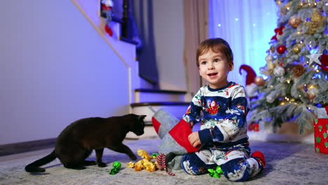 cute caucasian toddler and black cat sit on the floor. happy kid pulls the sweets from the stocking.