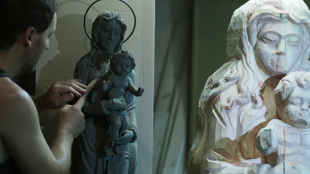 Artist meticulously sculpts religious Madonna and child statue with clay and wood carving tools in workshop, static camera shot