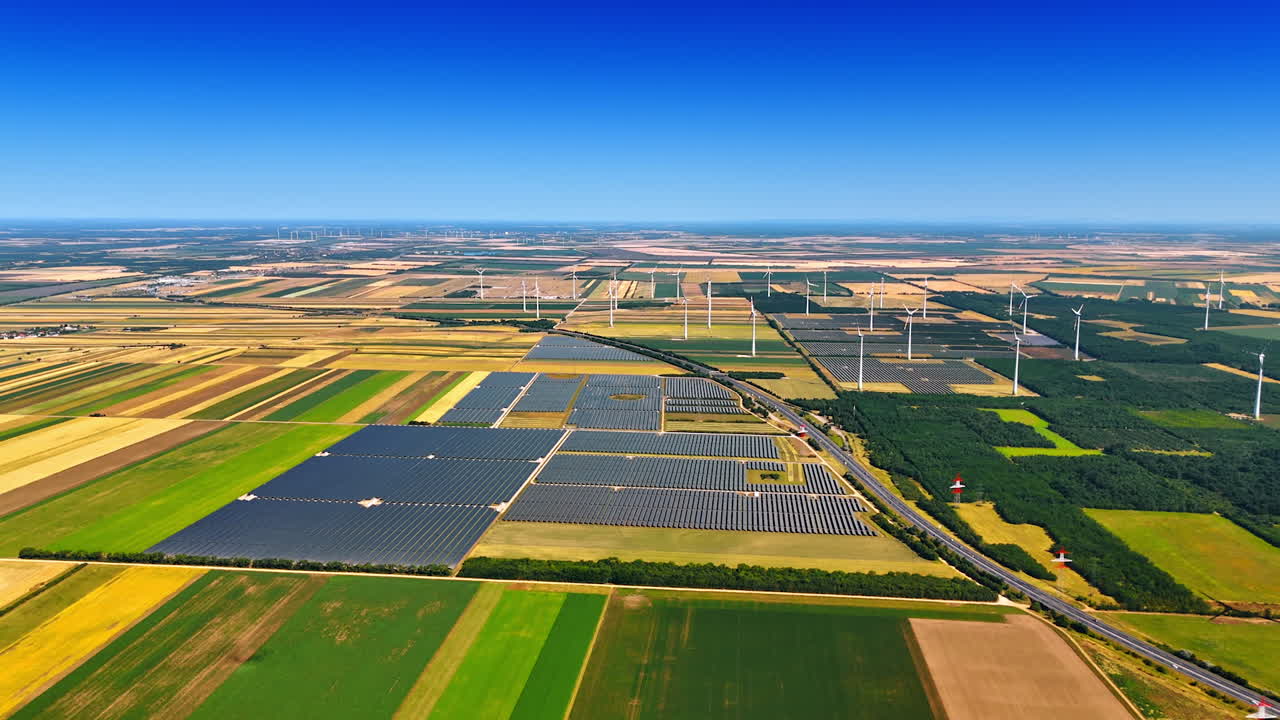 Fields with solar panels installed in the countryside. Wind farms in the rural area at backdrop. Aerial view