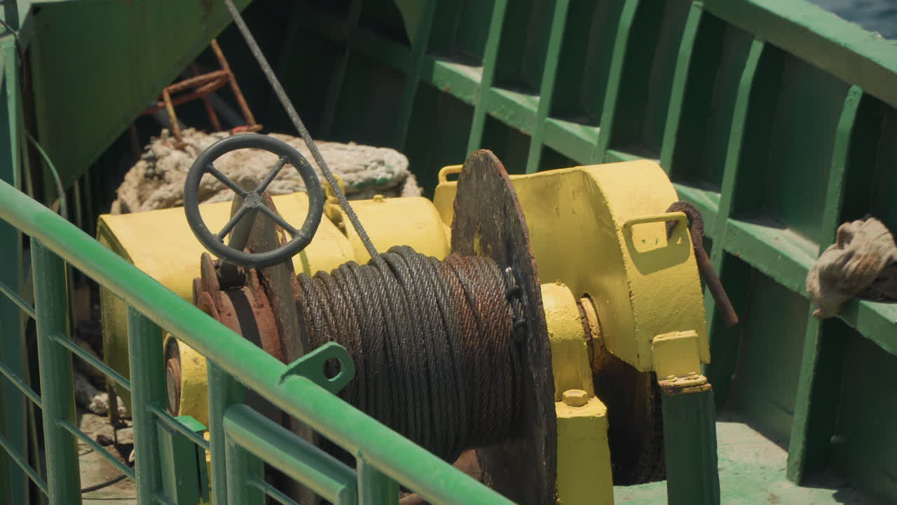 Cabrestante amarillo en cubierta de barco, carrete de cable oxidado y manivela, barandillas verdes enmarcando maquinaria pesada, escenario de mantenimiento con herramientas de ingeniero, pintura y texturas metálicas desgastadas por el sol, primer plano.