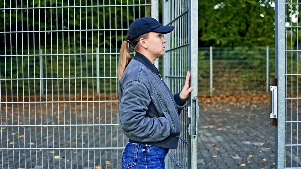 Una donna in giacca e berretto si trova vicino a un campo sportivo recintato con una rete. Una donna cupa sta dietro le sbarre. Una donna paranoica cammina in autunno.