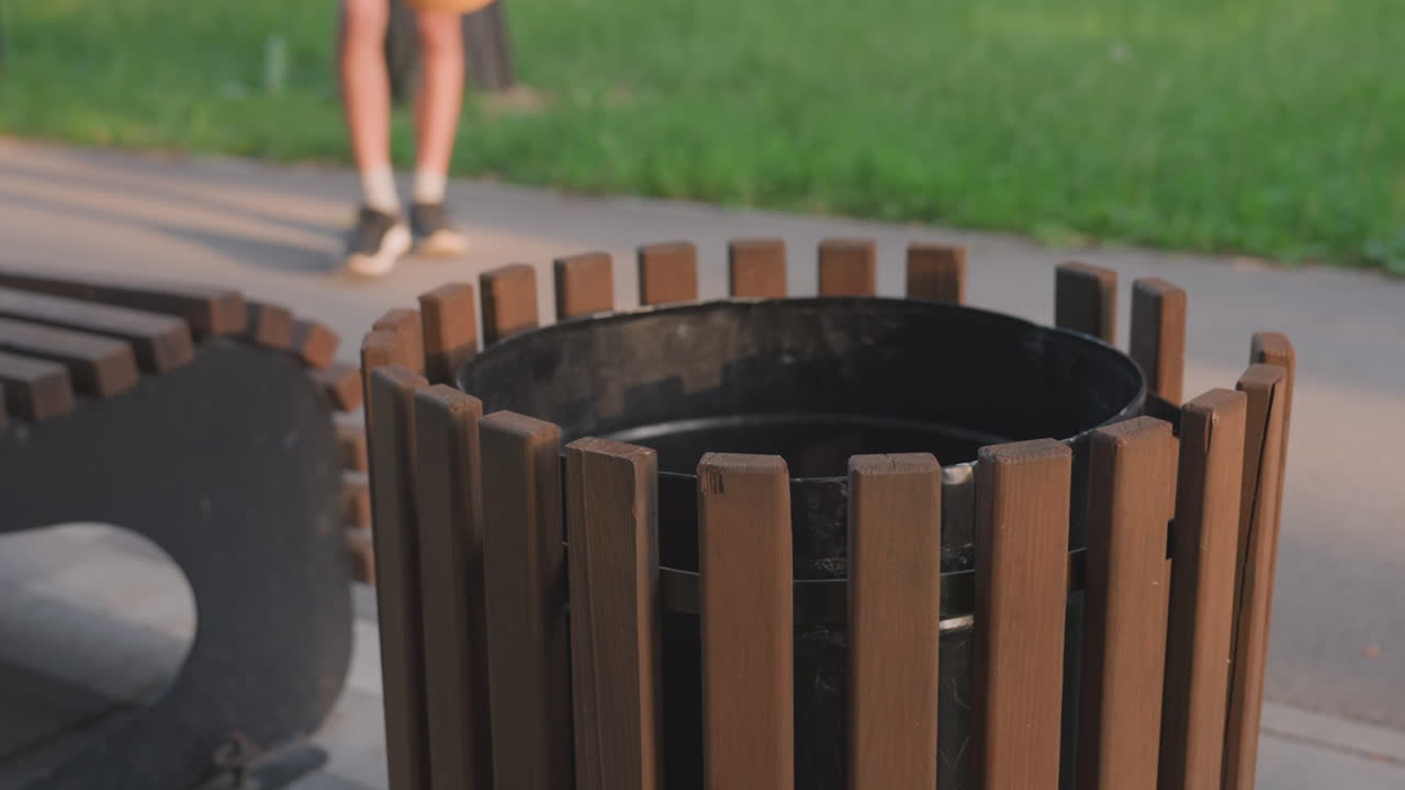 Park Passerby Hesitates Beside Bin, Young Adult In Yellow Shorts With Hand Hovering Over Wooden Waste Bin, Closeup On Legs And Hand, Urban Walkway And Green Grass Background, Tense Atmosphere