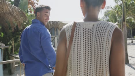 rear view of happy diverse couple talking, walking on sunny beach, in slow motion