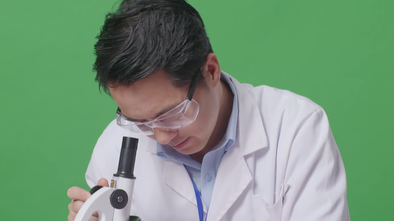 Close Up Of Asian Man Scientist Looking Through A Microscope On The Table With Test Tube Then Smiling And Crossing Her Arms While Sitting In The Green Screen Background Laboratory