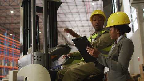 african american male and female workers wearing safety suits and talking in warehouse