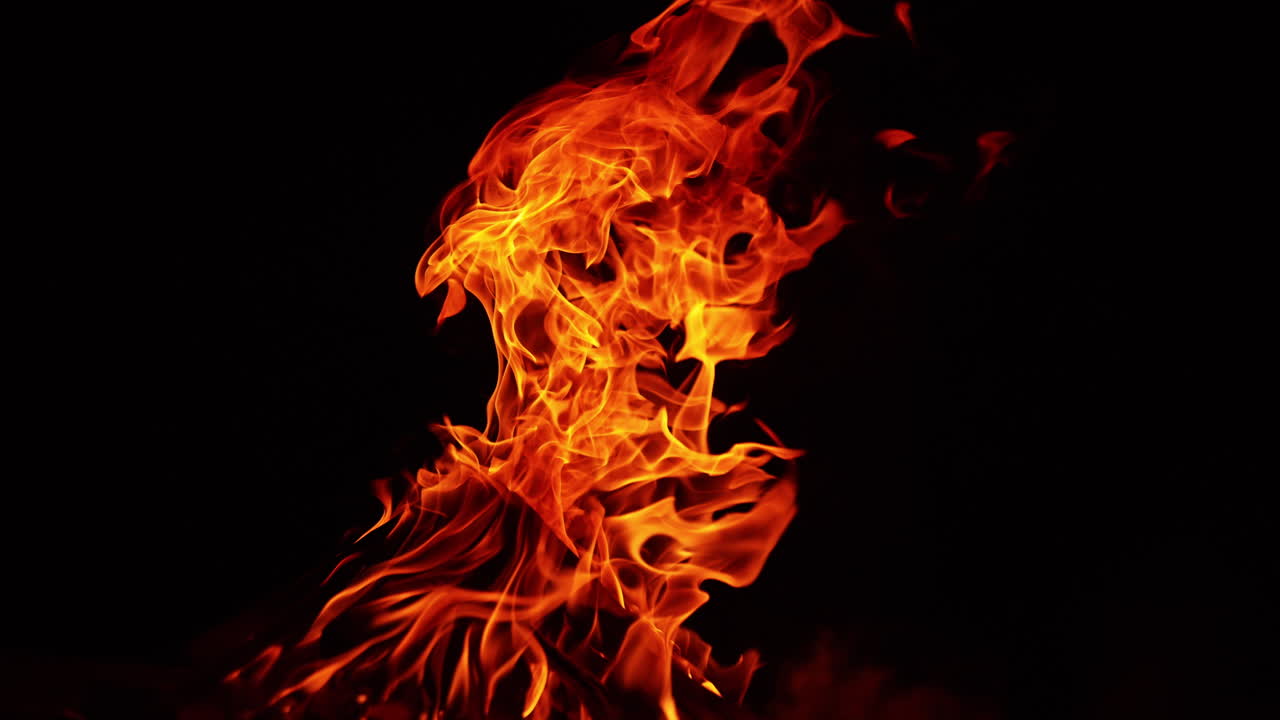 Wooden logs stacked in a pile burn in a big bonfire. Fire flames bending to the right in darkness.