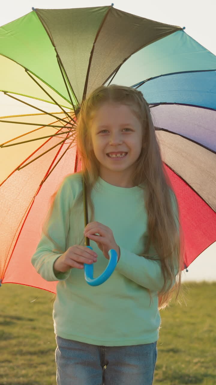 Girl swirls colorful umbrella against river. Parasol catches sunlight casting highlights of colors across rain-kissed landscape. Innocence of childhood