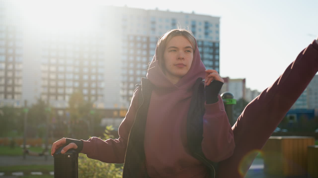 Fit lifestyle advocate holds pull up bar while stretching side leg high in urban outdoor park under bright sun, hoodie outfit, resilience and flexibility highlighted during dynamic fitness session