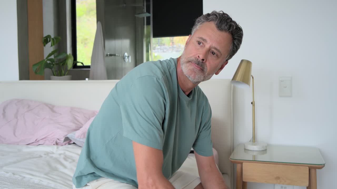 Uomo di mezza età reagisce a un rumore dal bagno, seduto sul letto in camera da letto, cercando la fonte.