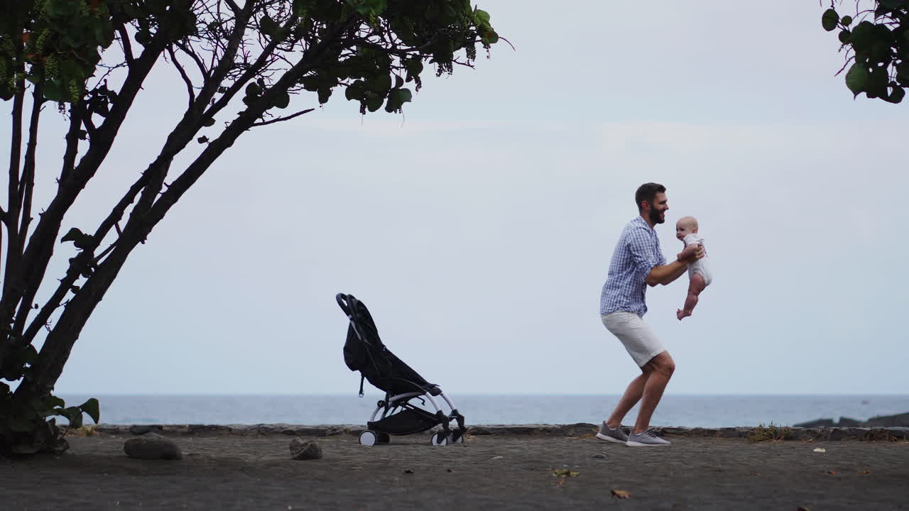 Against the sea backdrop, a portrait freezes a father's gentle kiss on his baby cheek. The vacationing family shows joy, dad embracing the toddler lovingly