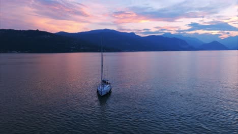 Eine-Ruhige-Luftaufnahme-Eines-Segelboots-Auf-Ruhigem-Wasser-Mit-Einem-Majestätischen-Sonnenuntergang-über-Den-Bergen-Im-Hintergrund