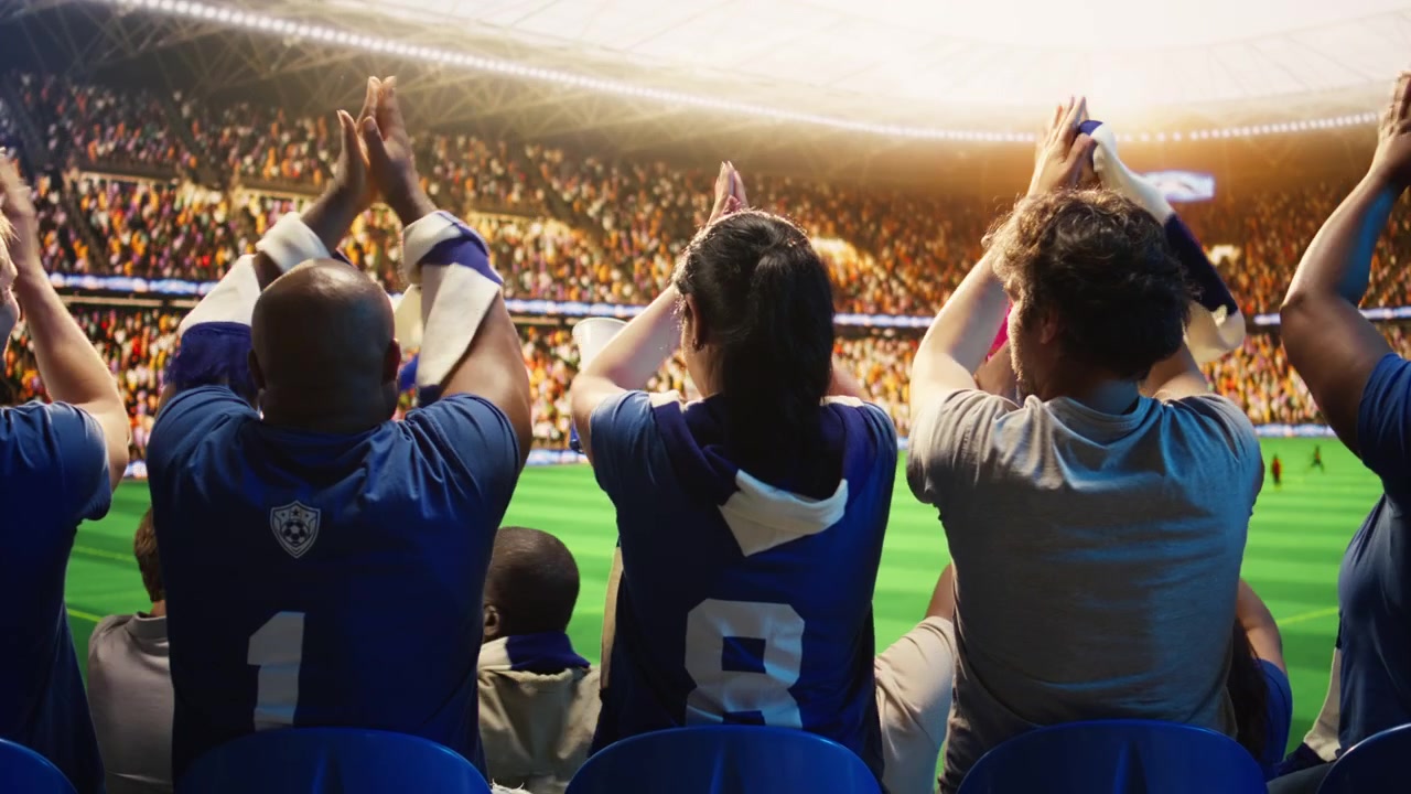 Aficionados al fútbol aplaudiendo al unísono desde los asientos del estadio durante un partido de fútbol en vivo.