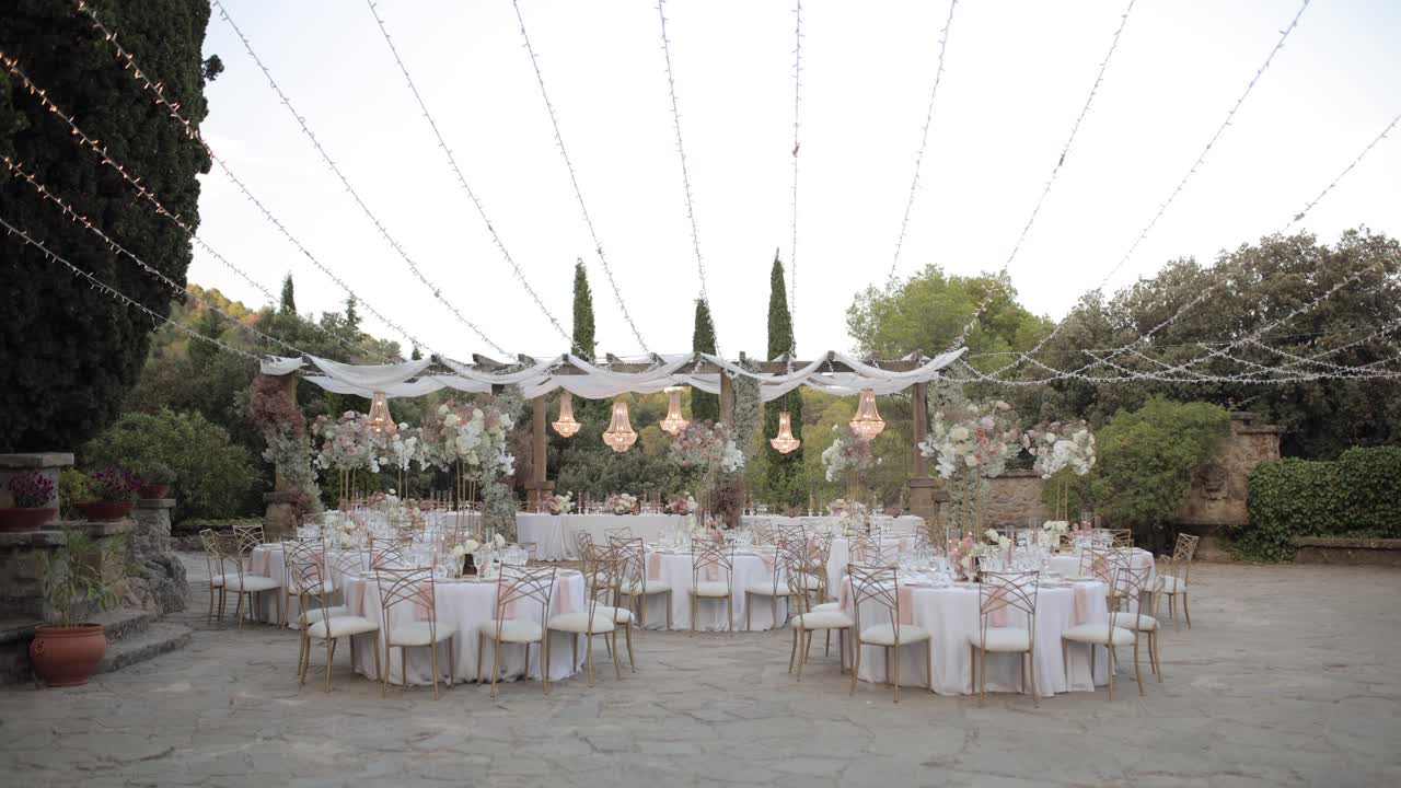 Allestimento elegante della tavola per una cena di matrimonio di lusso all'aperto con centrotavola floreali, candele e dettagli dorati. Atmosfera sofisticata con illuminazione romantica sotto un pergolato, perfetta per eventi di alto livello.