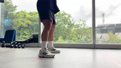 A person performs kettlebell swings in a gym with large windows, natural lighting, and a view of greenery