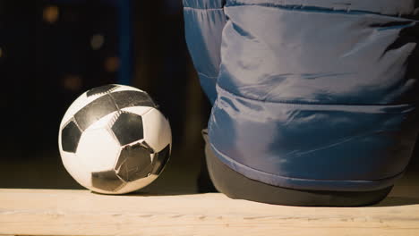 close up back view of a person in a blue hooded jacket seated on a wooden bench with a soccer ball by their side outdoors, under a spotlight in a dimly lit area