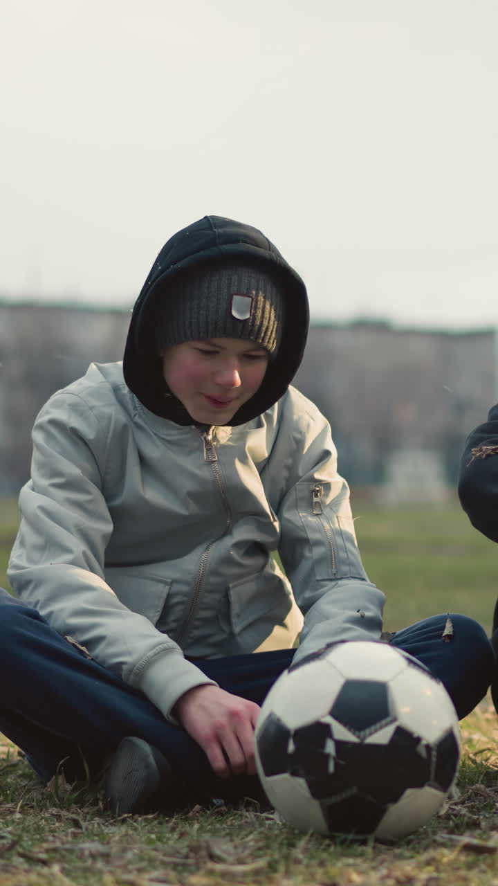 Two boys seated on a grassy field, engaged in discussion, with one in a gray top and a soccer ball in front of him, dry grass is scattered around them as they converse