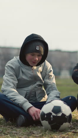 two boys seated on a grassy field, engaged in discussion, with one in a gray top and a soccer ball in front of him, dry grass is scattered around them as they converse