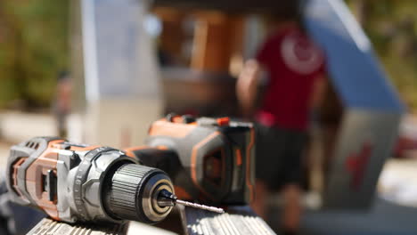 A-woodworker-places-a-cordless-drill-onto-a-bench-as-a-construction-worker-builds-a-custom-travel-trailer-on-a-job-site-in-the-background-with-copy-space-for-web-banner