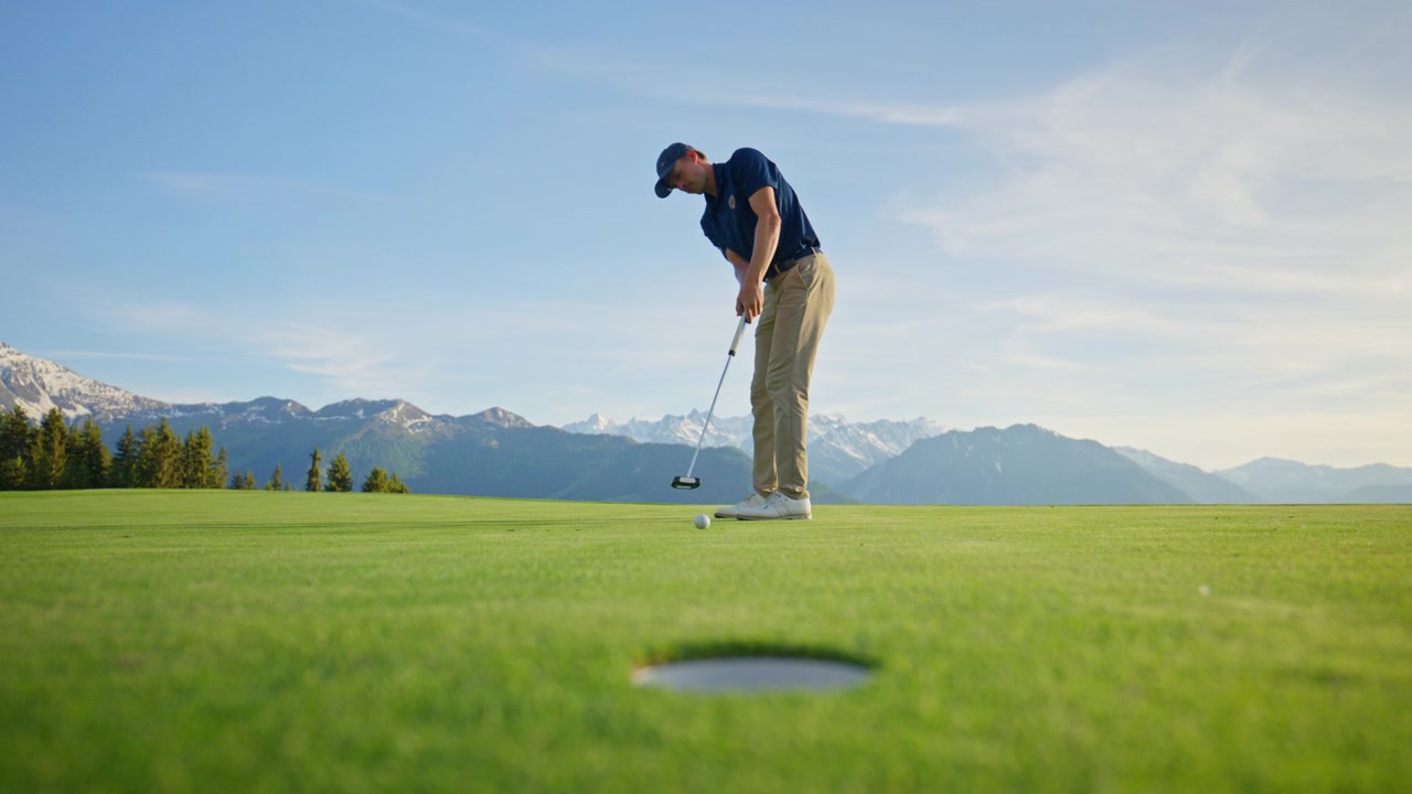 Inquadratura cinematografica dal basso di un golfista che tenta un putt su un campo da golf di montagna a Verbier, Svizzera. La pallina da golf rotola oltre la buca mentre un drammatico paesaggio alpino si estende sullo sfondo