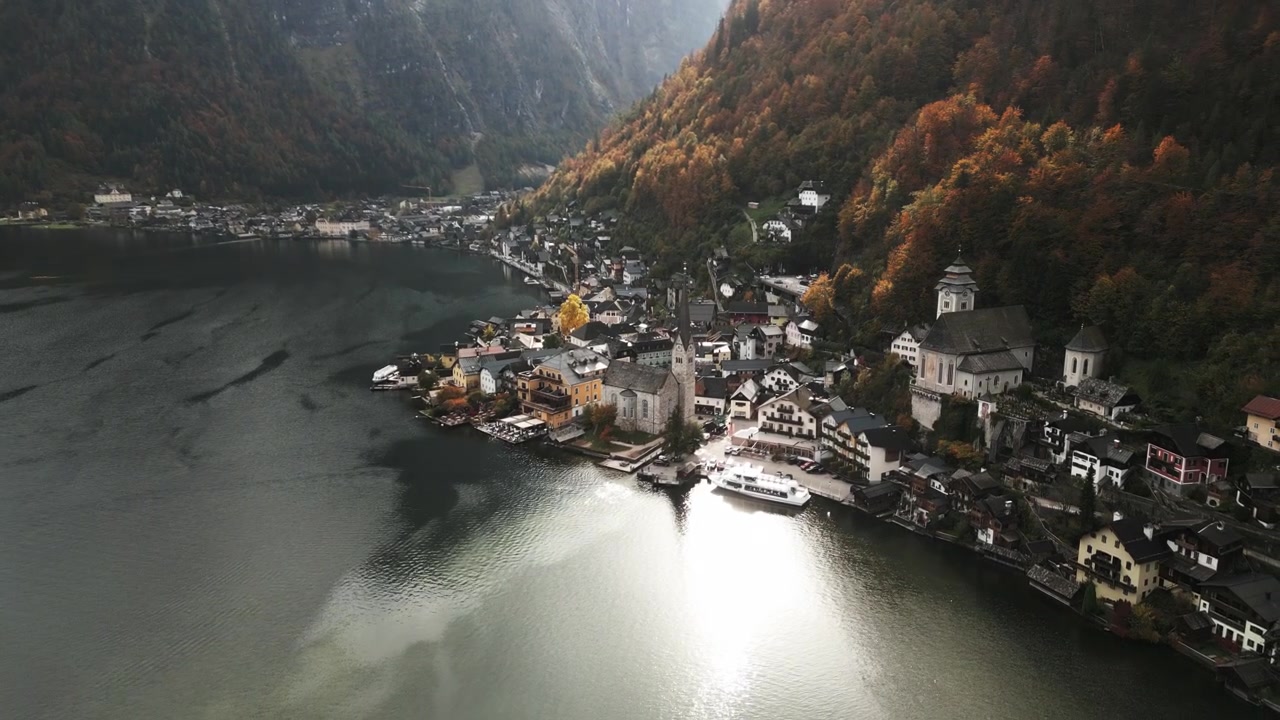 Med udsigt over en malerisk landsby ved søen, beliggende op ad stejle bjerge, fanger scenen levende efterårsløv og rolige vande, der reflekterer den charmerende arkitektur.