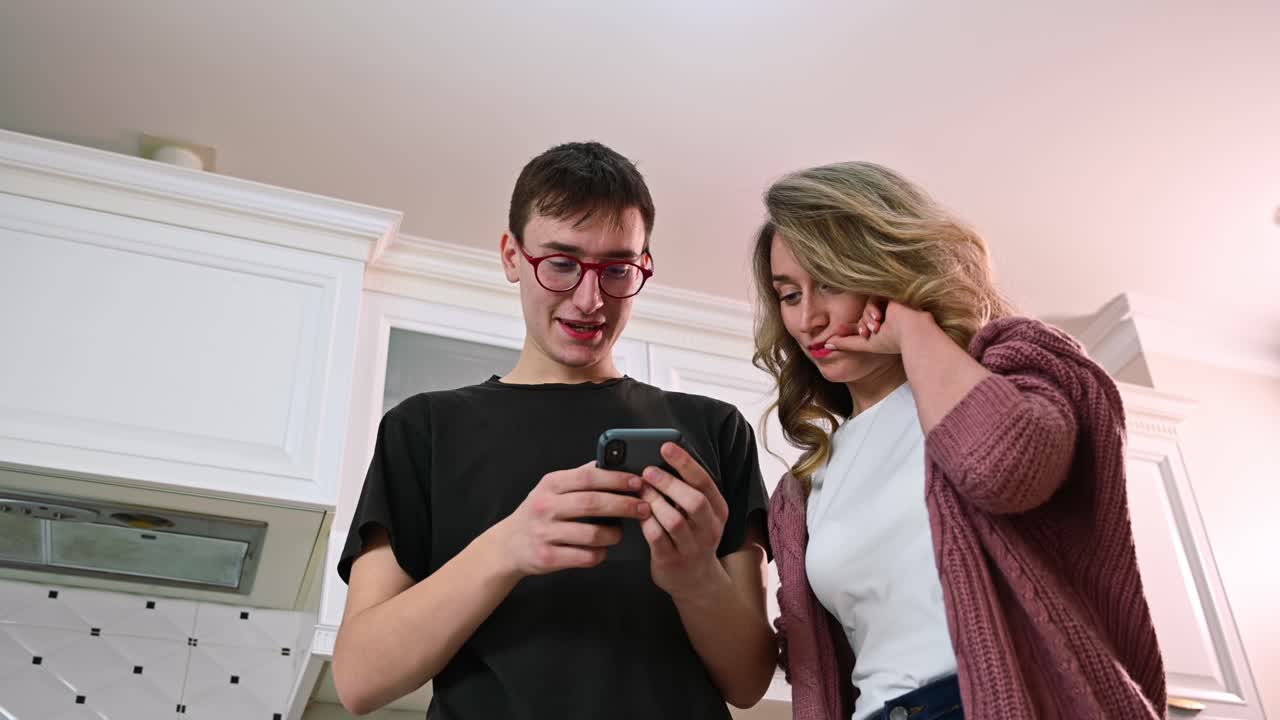 Young man and an interested woman looking at the phone in a white modern kitchen. Bottom view. Real emotions