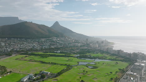 Fly-above-lush-green-lawn-on-golf-course-between-development-on-sea-coast.-High-and-flat-mountain-ridge-in-background.-Cape-Town,-South-Africa