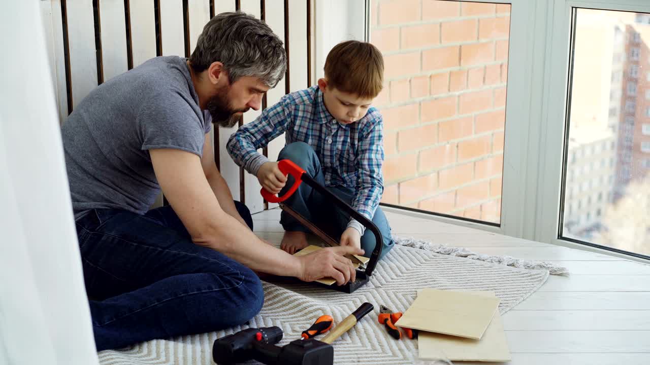 Little boy is focused on sawing piece of wood with hand saw with his father helping and teaching him. United family, construction work and childhood concept.