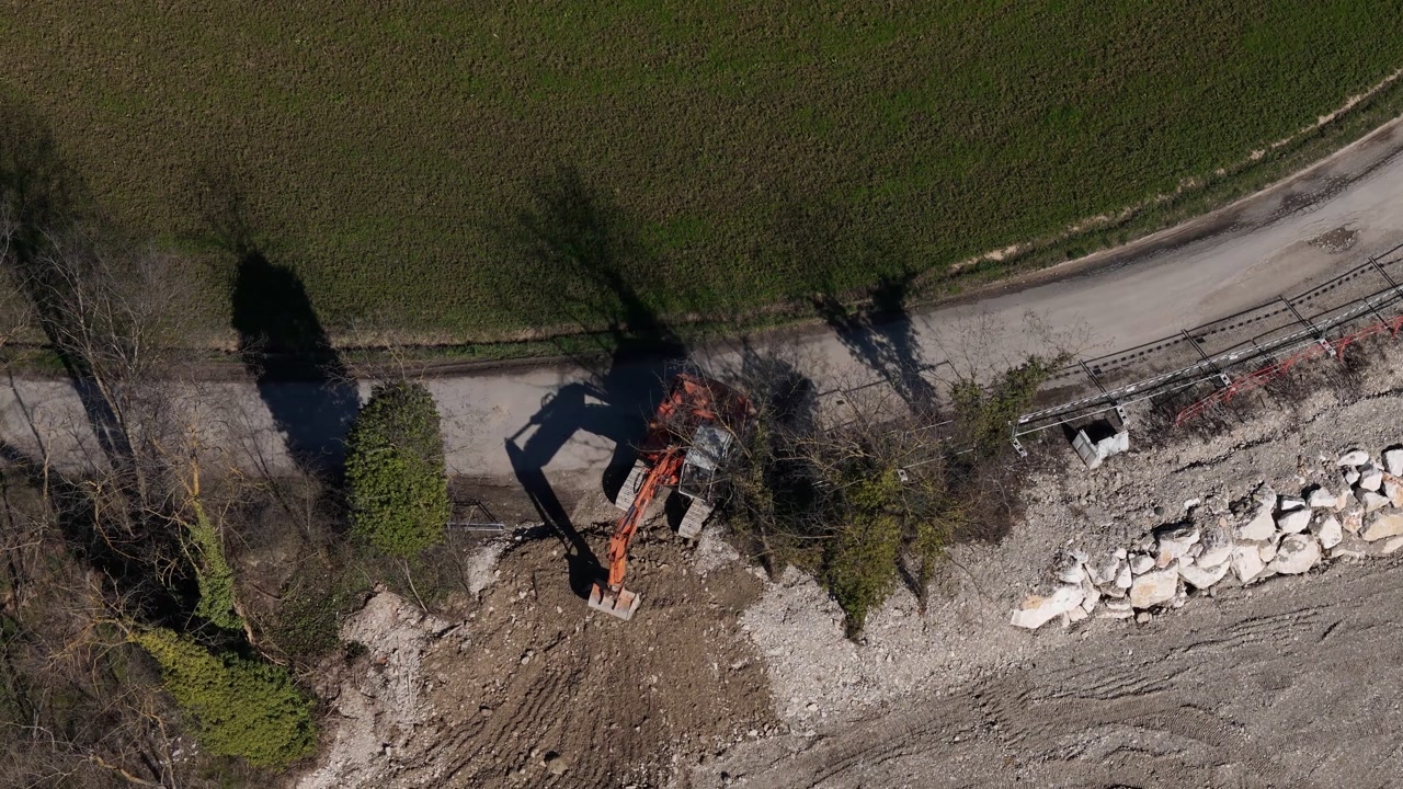 Drone footage captures an excavator at a construction site, surrounded by debris, rocks, and a dirt road, showcasing ongoing development