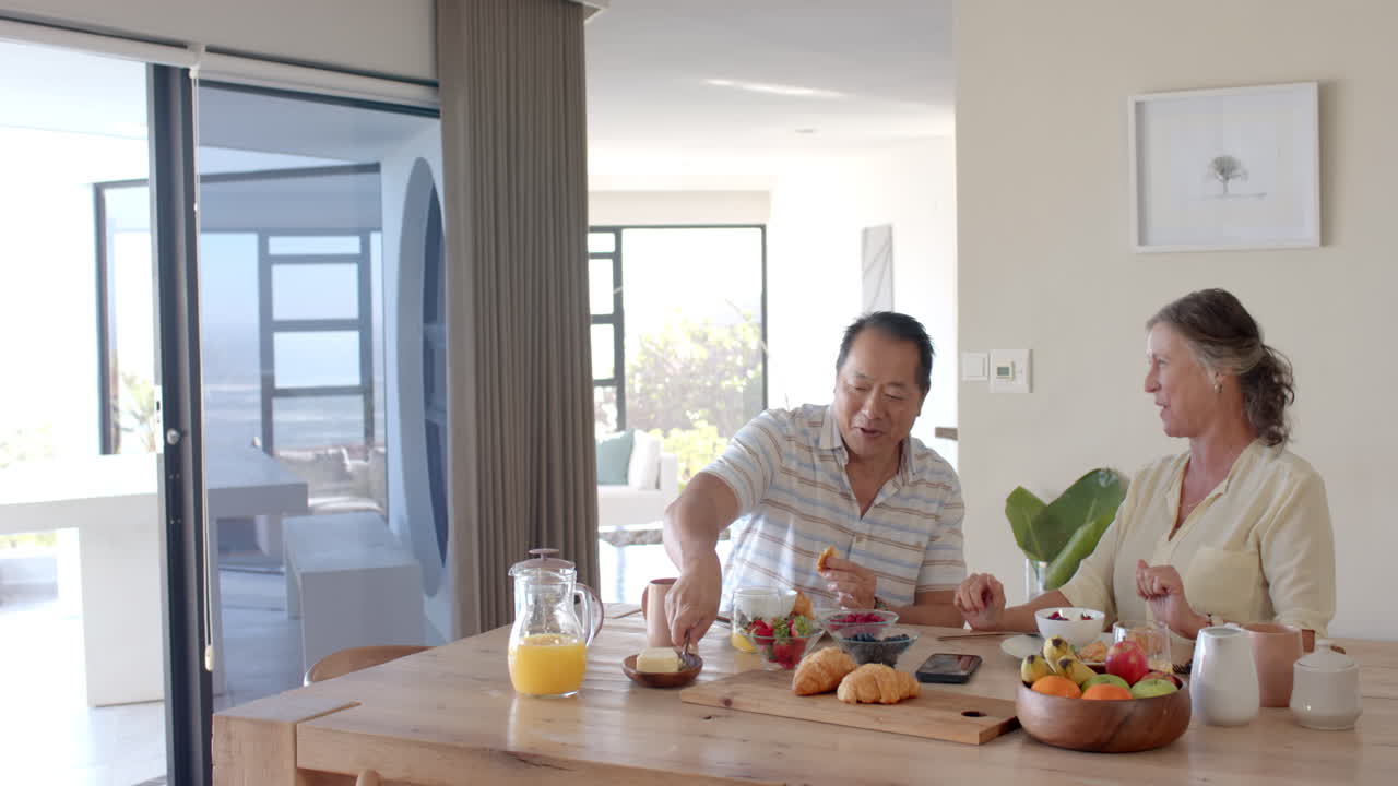 Having breakfast, senior couple enjoying fresh fruit and croissants at dining table