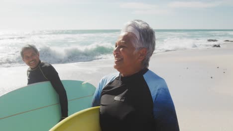 Happy-senior-hispanic-couple-walking-on-beach-with-surfboards