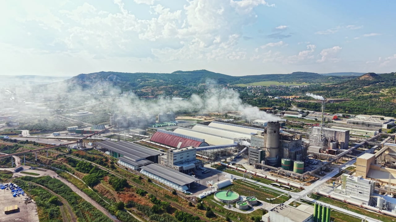 Industrial landscape showing a manufacturing plant with smoke rising from chimneys and machinery in operation near hills during the day