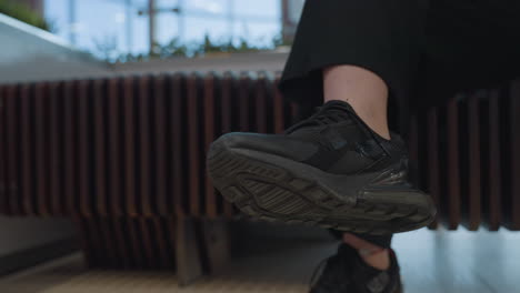close up of lady with crossed legs gently tapping foot to music while seated indoors. focus on black sneaker sole and rhythmic motion against artistic bench backdrop in modern mall environment