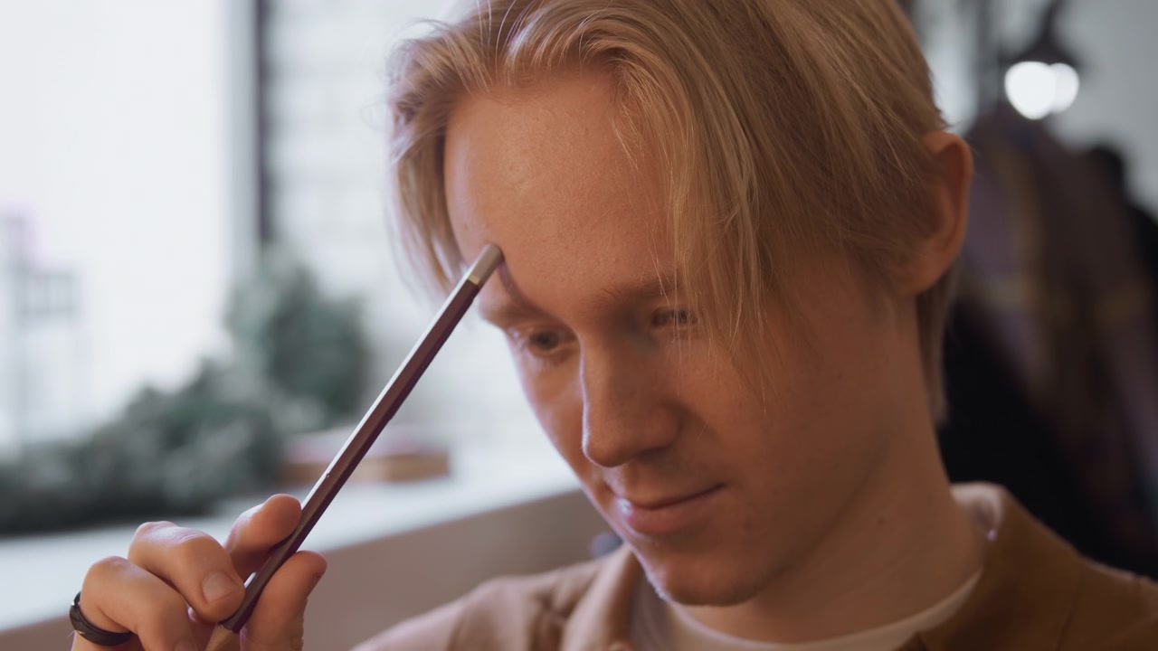 Primo piano di un giovane uomo che tiene ed esamina una matita in una luce naturale soffusa. La mano del soggetto, che indossa un anello sull'indice sinistro, afferra delicatamente lo strumento di scrittura in legno mentre il suo sguardo concentrato