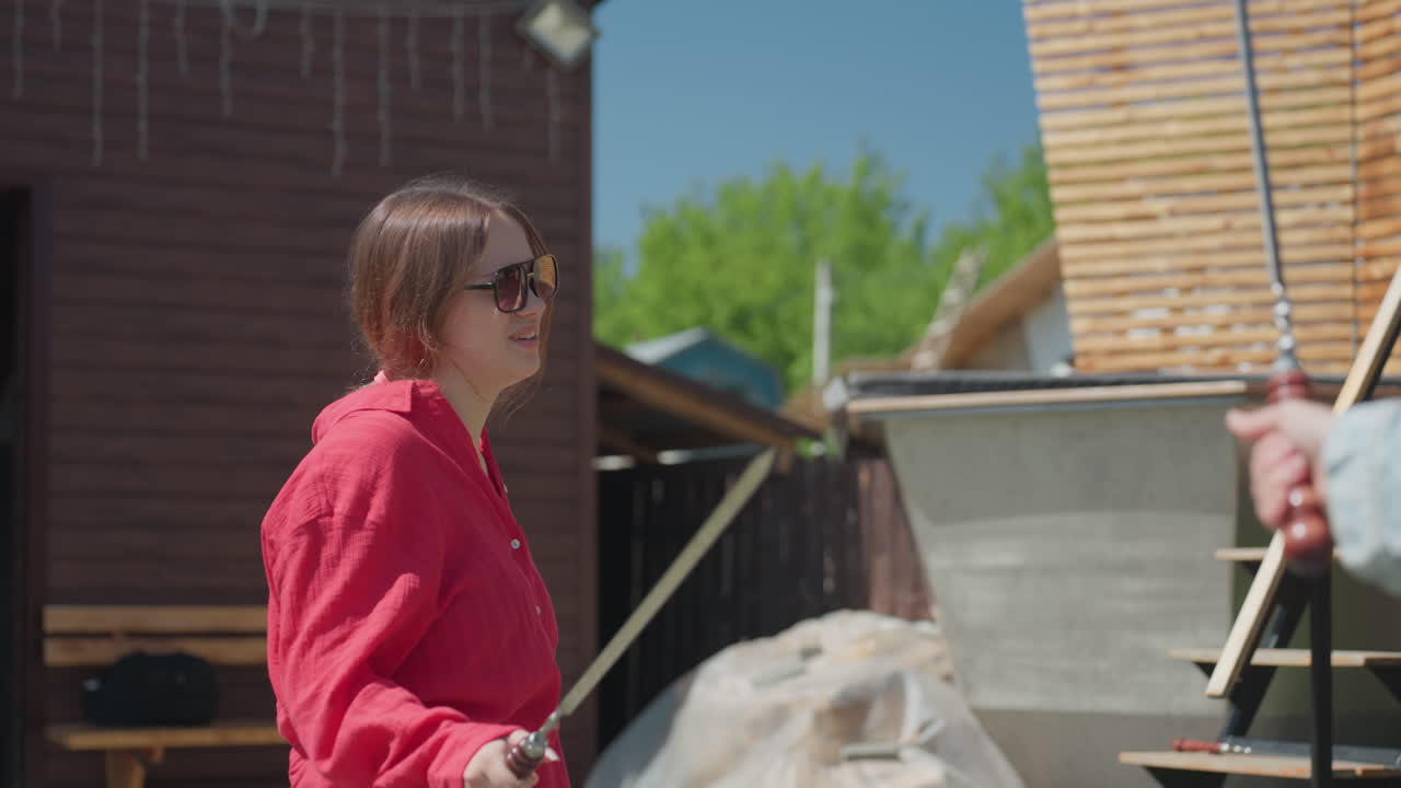 Confident woman in red shirt and sunglasses holds fencing sword while engaging in playful duel outdoors, standing near wooden structure under bright daylight with another sword raised in foreground