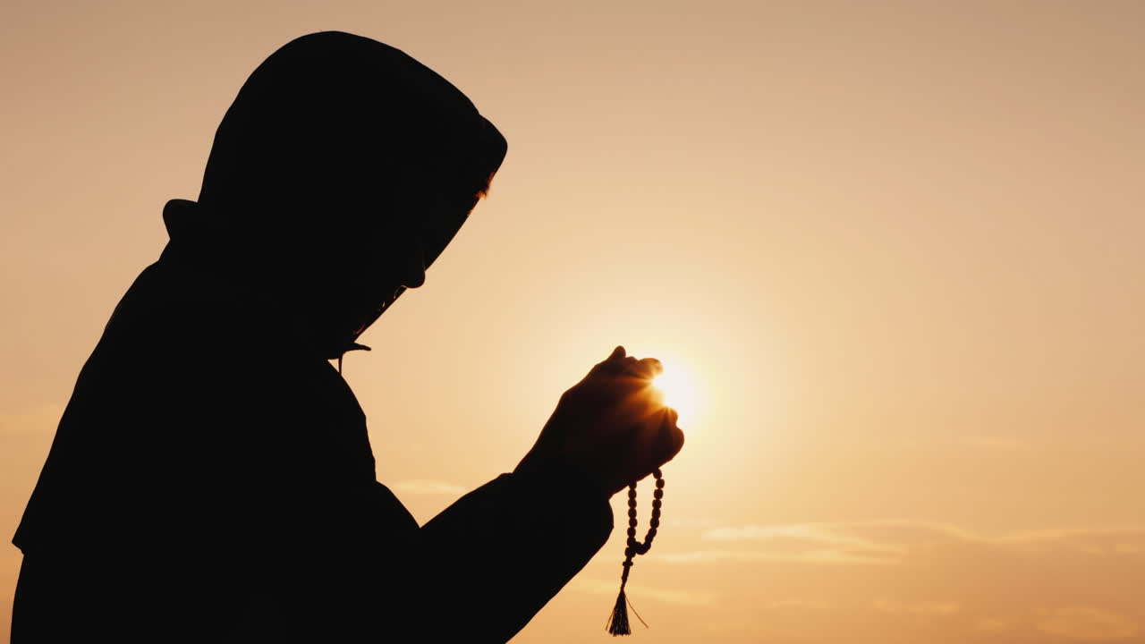 해질녘에 묵주를 통해 걸러내는 후드에 있는 남자의 실루엣