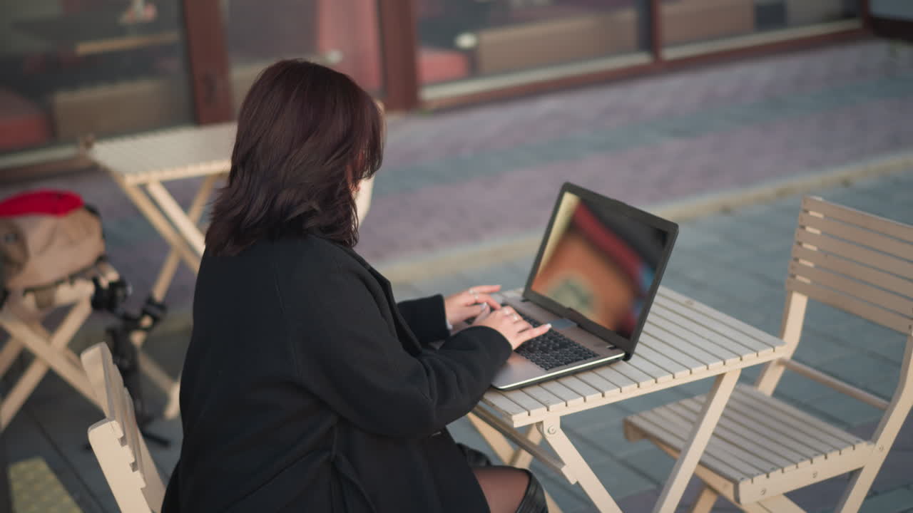 vista lateral de una mujer profesional escribiendo en una computadora portátil al aire libre en un entorno urbano, con un elegante abrigo negro, con muebles de madera blancos y arquitectura moderna en el fondo