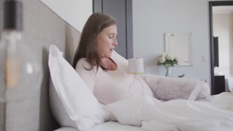 Woman-drinking-coffee-while-having-a-video-chat-on-her-laptop-in-bed-at-home