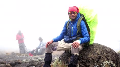 a black man sits and smiles with a backpack and jacket on a boulder during heavy fog