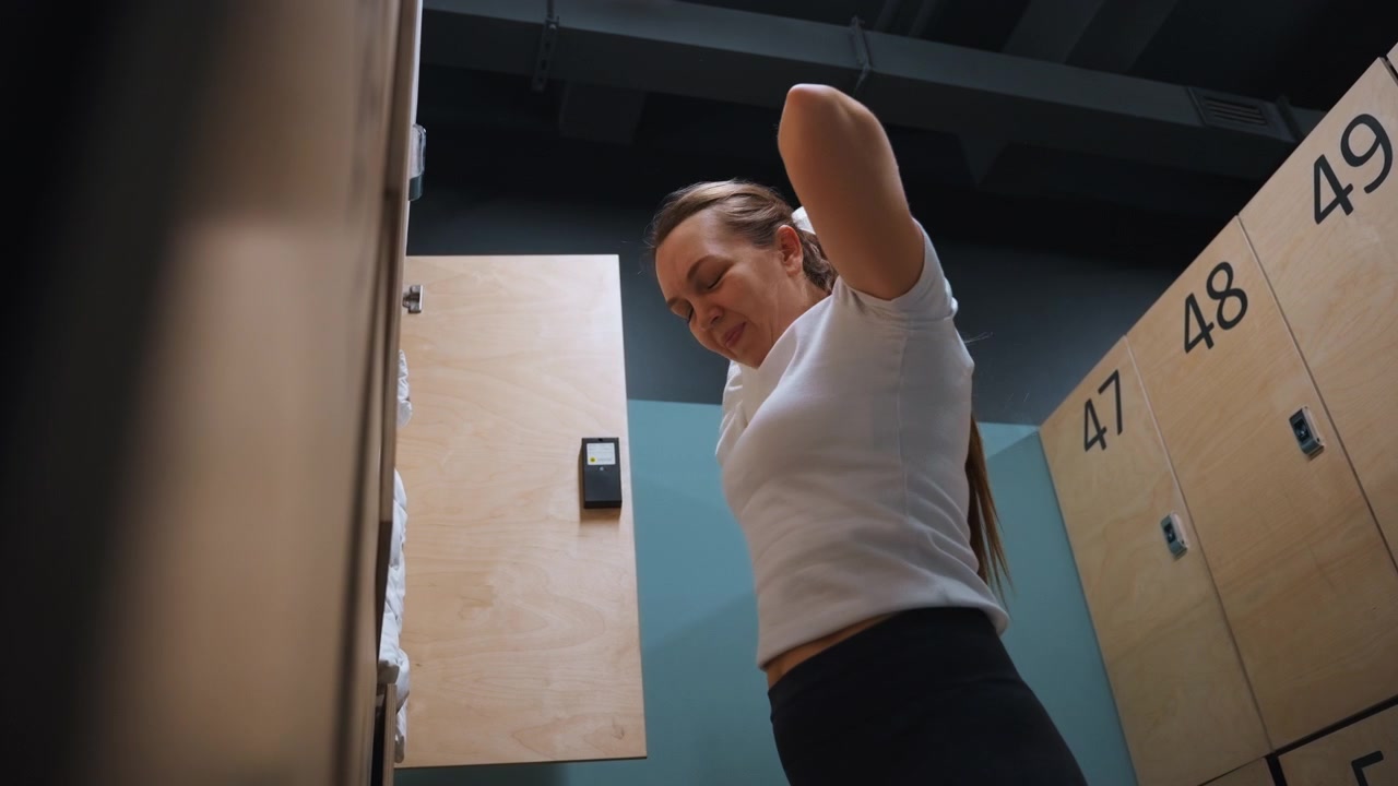 Atleta de badminton no vestiário preparando o equipamento, abrindo o armário e vestindo o moletom, ajustando a manga, armários de madeira numerados visíveis, iluminação fluorescente fria, rotina pré-jogo focada