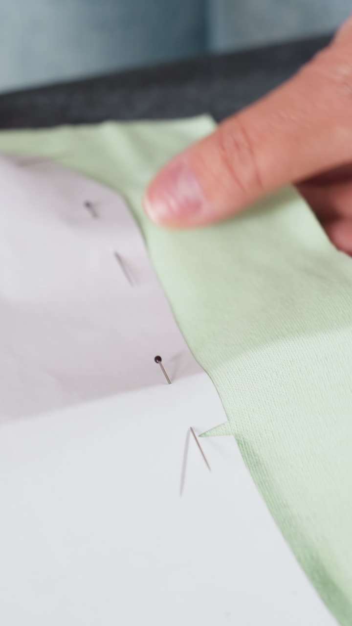 Dedos marcando muescas y revisando alfileres en un patrón de papel sobre tela verde; un primer plano revela grapas y pequeños cortes, una inspección meticulosa de pre-costura en una mesa con un ambiente de artesanía atento.