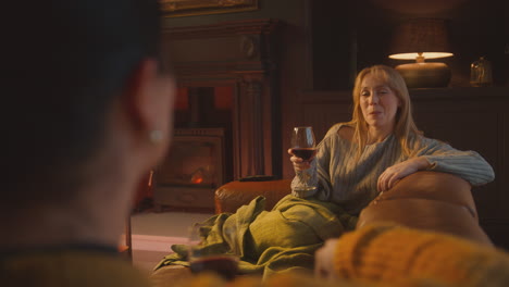 two female friends relaxing and laughing on sofa in lounge with cosy fire holding glass of wine