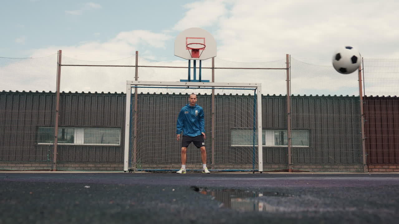 porta da calcio urbana bagnata, un giocatore solitario davanti a una recinzione metallica, pallone sovradimensionato in primo piano, riflessi di pozzanghere sull'asfalto, cielo nuvoloso, calci lenti e misurati e concentrazione silenziosa durante l'allenamento in solitaria