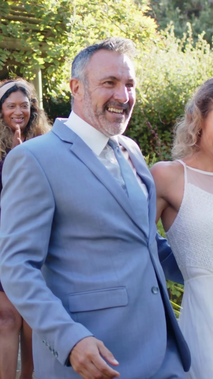 Video verticale: Sposo che ride e si gira dopo l'applauso di un invitato a un matrimonio, mostrando abito blu e vestito.