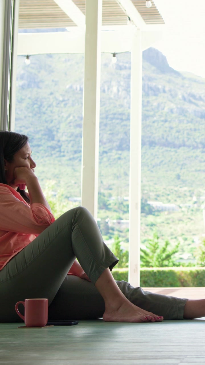 Video verticale: Donna in abito color corallo a casa che posiziona il telefono accanto a una tazza, si sposta e guarda la montagna, con spazio per il testo.