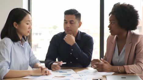 collaborative meeting among three professionals analyzing data and discussing strategies in a bright office setting with documents and charts spread on the table