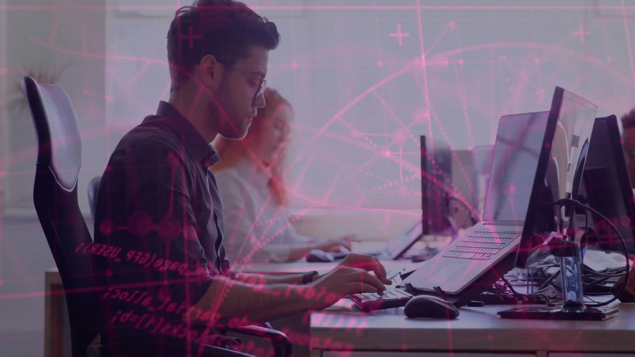 Male software developer typing on keyboard at open-plan tech office monitors with pink data overlay