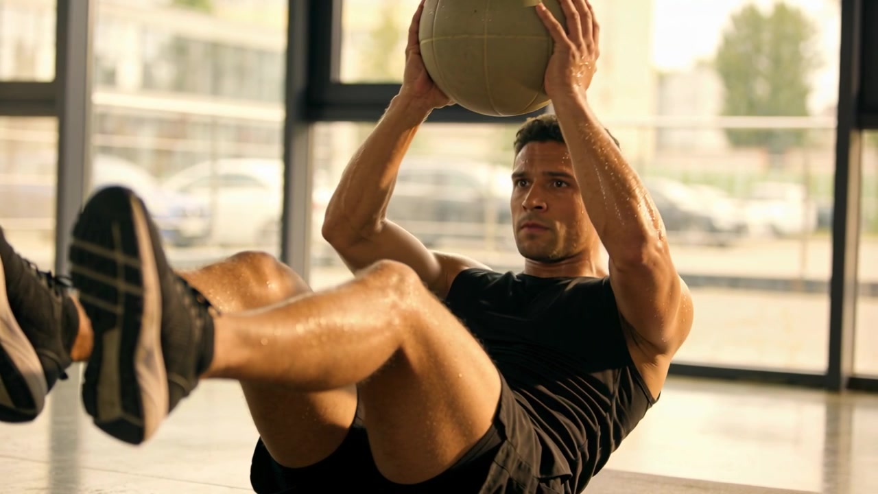 Inizio serie, uomo in pantaloncini neri che esegue un V-sit su un tappetino in palestra, sollevando una palla medica per gli addominali